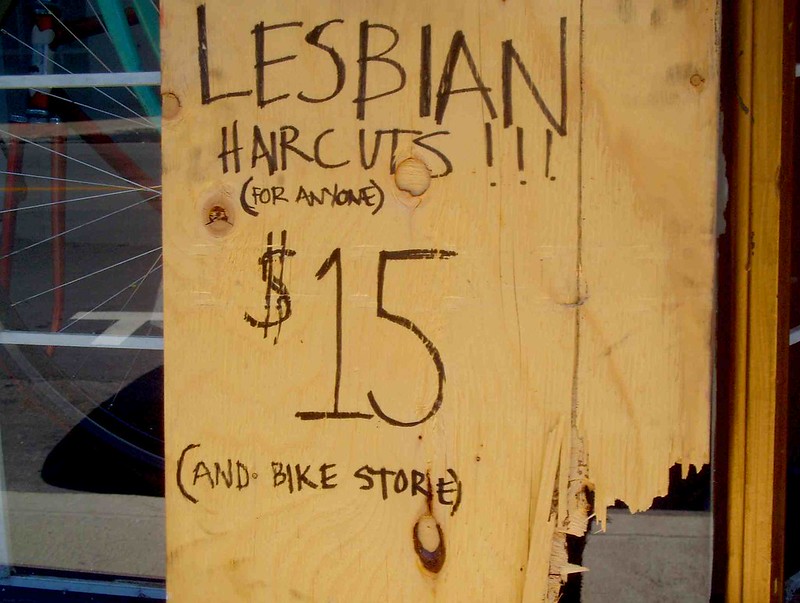 A wooden sign with Lesbian Haircuts!!! (for anyone) $15 (and bike store) written on it with black pen in the window of a bike store.